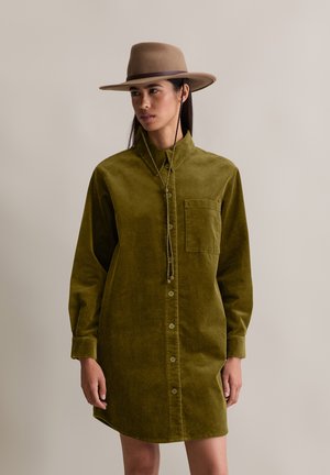 Olijfgroene corduroy jurk met een gestructureerde kraag, lange mouwen, borstzak en knoopsluiting aan de voorkant. Draag het met een tan brede hoed.