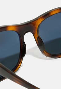 Ochelari de soare tortoise brown cu lentile întunecate și o textură netedă. Au un arc curbat pe sprânceană și un design elegant și contemporan al ramei.