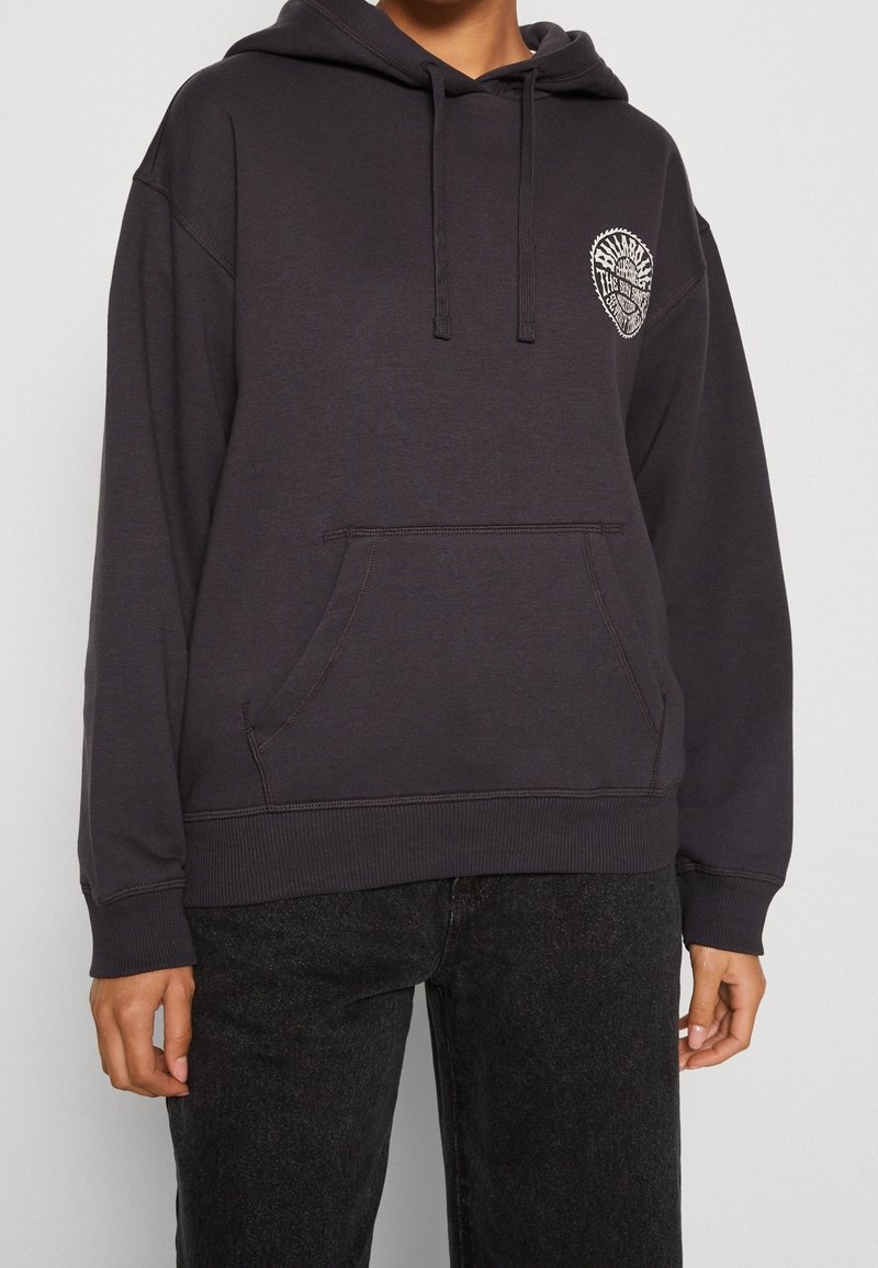 Osoba oblečená v černé mikině s kapucí a přední kapsou, na které je malé bílé kruhové logo na levé hrudi, v kombinaci s černými džíny.