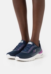 Zapatillas de running en navy con acentos morados, parte superior de malla, suela blanca, patrón de rayas, lengüeta acolchada y logotipo en el lado y el talón.