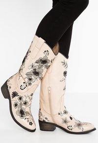 Stivali in pelle beige alti fino al ginocchio con ricami floreali neri, una cerniera laterale e un tacco in legno impilato. Texture liscia e design strutturato.