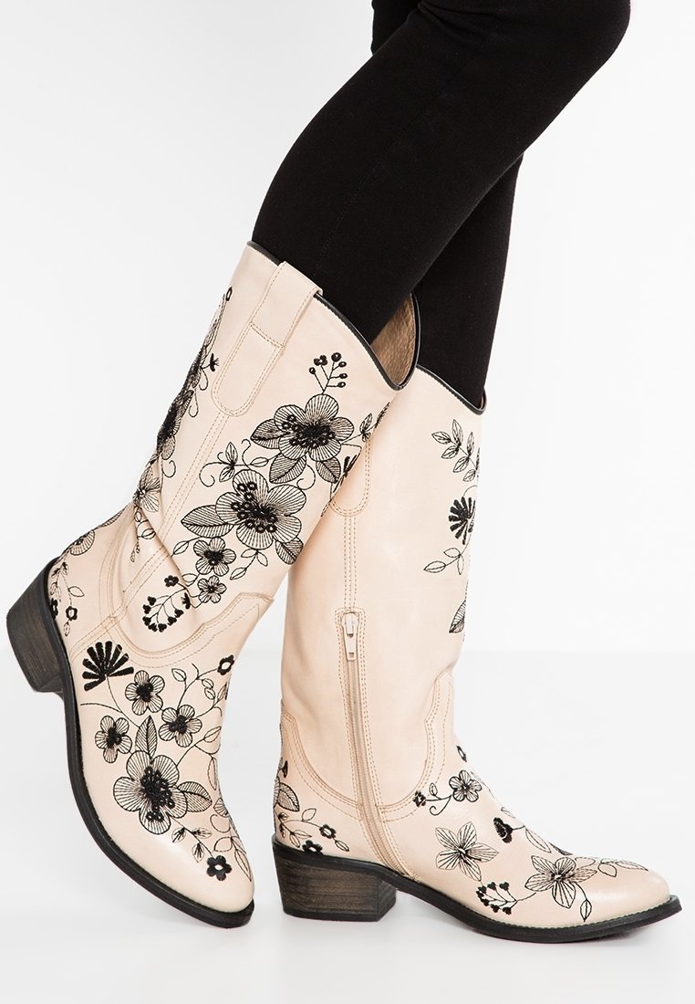 Stivali in pelle beige alti fino al ginocchio con ricami floreali neri, una cerniera laterale e un tacco in legno impilato. Texture liscia e design strutturato.