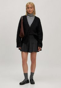 Jonge vrouw met blond haar, gekleed in een gestreepte turtleneck, zwarte cardigan, donkere rok, grijze sokken, zwarte schoenen en een bruine schoudertas.
