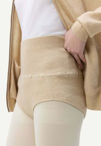 Höga beiga stickade shorts har en ribbad textur, en tajt passform och är kombinerade med genomskinliga ljusa strumpbyxor och en vit topp.