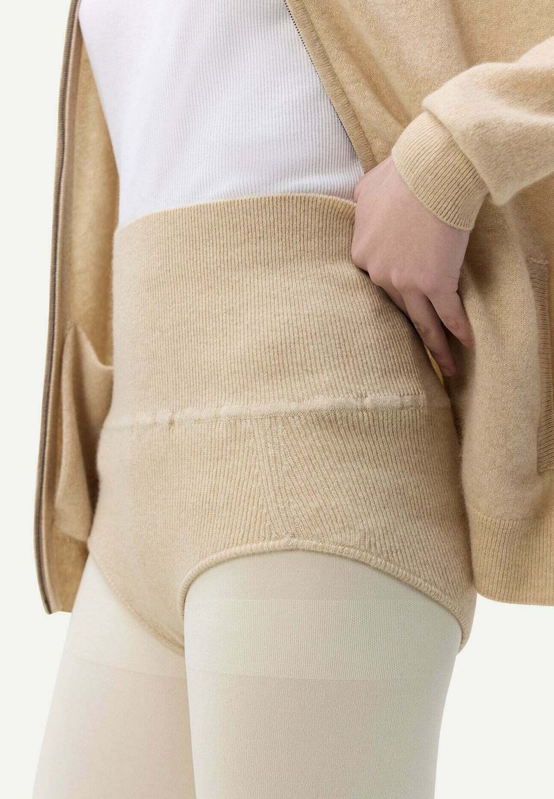 Höga beiga stickade shorts har en ribbad textur, en tajt passform och är kombinerade med genomskinliga ljusa strumpbyxor och en vit topp.