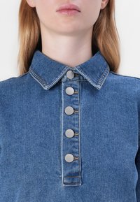 Denimskjorte med en strukturert krage, som har fem metallknapper og synlige sømdetaljer i en lys blå nyanse.