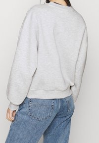 Felpa grigia chiara con vestibilità rilassata, polsini e orlo a coste, abbinata a jeans blu a vita alta. Tessuto testurizzato, cuciture visibili.