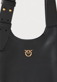 Bolsa de couro preto com uma textura suave, apresentando um logótipo de abelha dupla em tom dourado e alças acentuadas com tachas douradas.