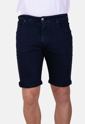Hombre que lleva puestos unos shorts de mezclilla azul oscuro remangados en el dobladillo, combinados con una camisa blanca metida dentro, visible desde la cintura hasta por encima de las rodillas.