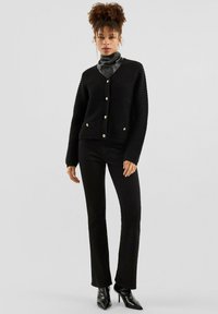 Frau mit lockigem Haar, die eine schwarze strukturierte Strickjacke, schwarze Hosen, schwarze spitze Stiefel mit Absatz und einen schwarzen Lederhalstuch trägt.