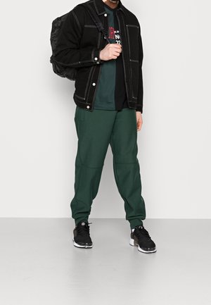 Black denim jacket with white stitching, black graphic t-shirt, green jogger pants, and black sneakers, carrying a black backpack.