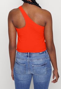 Oranžový žebrovaný crop top s asymetrickým designem, kombinovaný s světle modrými džíny s kapsami na zadní straně a jemným nažloutlým detailem.
