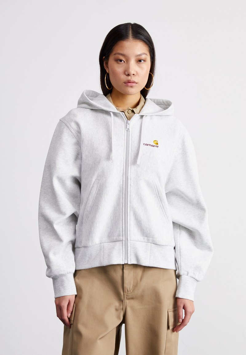 Grå zip-up hettegenser med en tilpasset midje, stor hette og frontlommer. Carhartt-logoen er brodert i gult og mørkerødt.