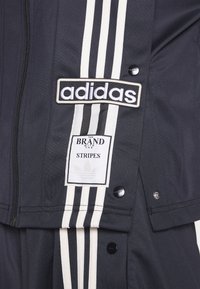 Marinträningsdräkt med vita diagonala ränder, en framträdande Adidas-logotyp och en etikett som säger "VARUMÄRKET MED DE TRE RÄNDERNA."