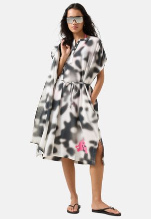 Kvinde iført en løs grå og sort tie-dye kjole med en pink logo, solbriller og sorte flip-flops, står med den ene hånd i lommen.