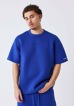 Jeune homme aux cheveux bouclés portant un haut bleu à manches courtes et un pantalon bleu assorti, debout sur un fond blanc uni.