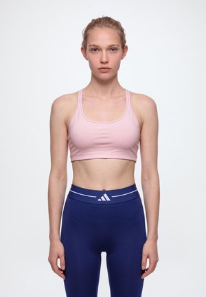 Une femme se tient face à l’objectif sur un fond uni, vêtue d’un soutien-gorge de sport rose et d’un legging bleu marine.