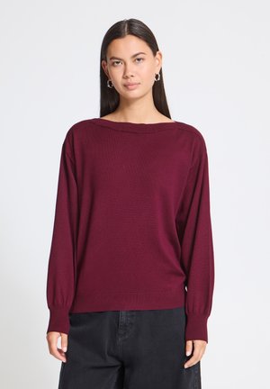Femme aux longs cheveux foncés portant des boucles d'oreilles créoles argentées et un pull bordeaux ample avec un pantalon noir sur un fond uni.