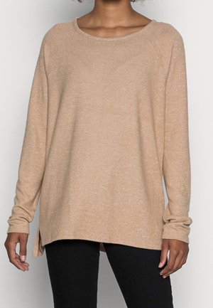 Personne portant un pull beige à manches longues en maille avec des fentes latérales et un pantalon noir, debout devant un fond uni.