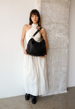 Femme en robe blanche et bottes noires tenant un grand sac en cuir noir à large bandoulière, debout contre un mur en béton texturé.
