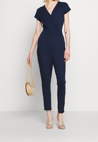 Granatowy kombinezon z krótkim rękawem, dekoltem w serek i dopasowanym w talii. Posiada zwężane nogawki oraz teksturowaną tkaninę. W połączeniu z małą, lekką torbą.