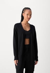 Cardigan long noir en tissu doux. Il présente un décolleté en V, une ouverture sur le devant et deux poches latérales, avec des poignets et un ourlet côtelés.