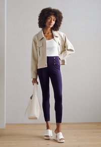 Beige cropped jack, witte fitted top, hoge taille marineblauwe leggings met knopen, witte blokhak sandaaltjes, draagt een witte tas.
