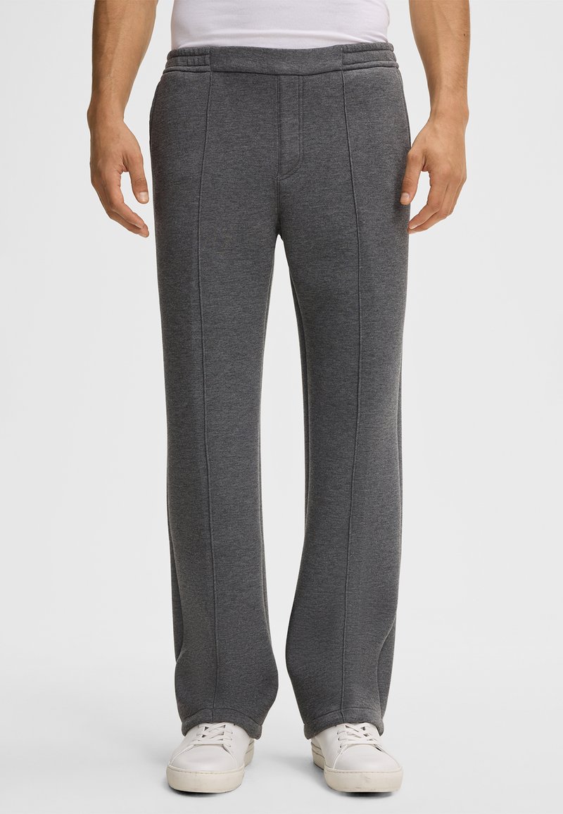 Mann trägt dunkelgraue, gerade geschnittene Sweatpants und weiße Sneaker, steht vor einem schlichten weißen Hintergrund.