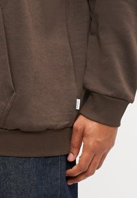 Brun hette laget av myk stoff med ribbestrikkede mansjetter, kengurulomme og en liten merkeetikett på siden. Behagelig passform med en glatt tekstur.