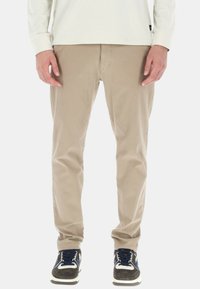 Pantaloni in cotone beige con una vestibilità dritta, caratterizzati da una chiusura con cerniera e passanti per cintura, abbinati a sneakers scure e a una maglietta bianca a maniche lunghe.