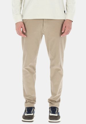 Pantaloni in cotone beige con una vestibilità dritta, caratterizzati da una chiusura con cerniera e passanti per cintura, abbinati a sneakers scure e a una maglietta bianca a maniche lunghe.