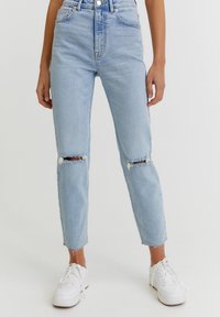 Vaqueros de tiro alto en azul claro con detalles de rodillas desgastadas, que presentan un diseño de pierna recta y bajos deshilachados, combinados con zapatillas blancas.