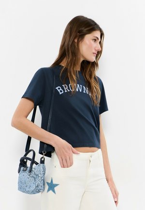 Mujer joven con una camiseta corta azul marino con el texto "BROWNIE", pantalones blancos con un parche de estrella azul y llevando un pequeño bolso azul con estampado.