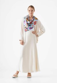Robe longue ivoire avec une silhouette fluide, associée à un foulard en soie florale colorée. Mannequin portant des sandales en plastique blanches décontractées.