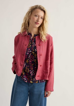 Jonge vrouw met golvend blond haar, gekleed in een rood jasje met knopen over een bloemenblouse en blauwe spijkerbroek, staand tegen een effen achtergrond.
