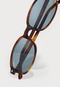 Ray-Ban aurinkolasit, joissa on kilpikonnakuosiset ruskeat suorakulmaiset kehykset ja tummat linssit, aseteltuna vaalealle pinnalle.
