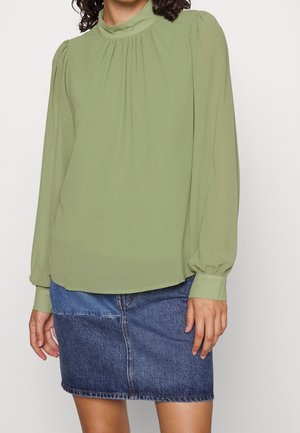 Kobieta ubrana w jasnobeżową bluzkę z długim rękawem z marszczonym dekoltem i dwukolorową mini spódniczkę dżinsową.