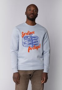 Sweatshirt bleu clair avec un graphique d'une boîte de sardines et du texte orange et bleu indiquant "Sardines Portugal." Tissu doux, design col rond.