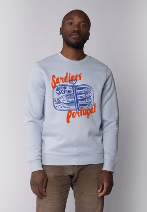 Lichtblauwe sweatshirt met een grafiek van een sardineblikje en oranje en blauwe tekst met de tekst "Sardines Portugal." Zachte stof, ronde halsontwerp.