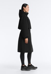 Femme debout de profil, portant un long manteau noir à capuche avec des poignets élastiques, des leggings noirs et des chaussures de sport noires, sur un fond blanc.