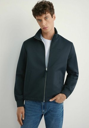 Granatowa kurtka na zamek z wysokim kołnierzem, ściągaczami na rękawach i kieszeniami bocznymi, noszona na białej koszulce. Gładka faktura tkaniny, nowoczesny design.