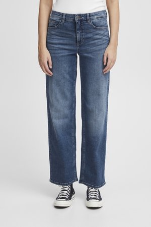 Personne portant un jean bleu taille moyenne à jambes larges avec un effet délavé et des baskets montantes noires, debout devant un fond uni.
