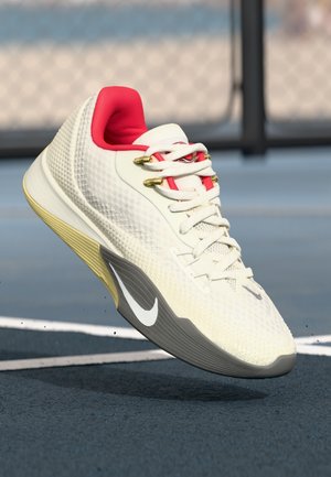 Biało-czerwony sneaker Nike w powietrzu nad ciemnoniebieskim kortem tenisowym, z widocznym bocznym logo i szczegółami fakturowanej podeszwy.