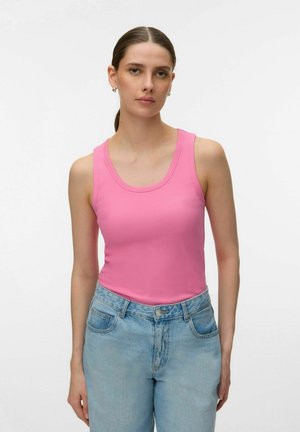 Young woman with brown hair in a pink sleeveless top and light blue jeans standing against a plain white background.