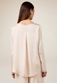 Langärmlige Bluse aus weichem, hellem Beige-Stoff, mit Rundhalsausschnitt, Rückenfalte und abgerundetem Saum. Glatte Textur, minimalistisches Design.