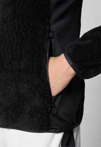 Jachetă neagră cu un corp din fleece texturat, mâneci din material neted, buzunar lateral cu fermoar și accente negre. Vedere de aproape a materialului și detaliilor.