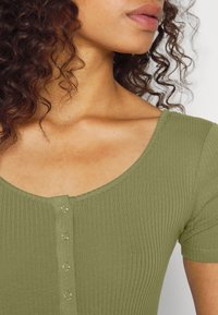 Zelenohnědý elastický top s krátkými rukávy, oválným výstřihem a pěti knoflíkovými zapínáními vpředu. Měkká textura a přiléhavý střih.