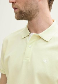 Polo shirt jaune clair avec un tissu texturé, patte de boutonnage à deux boutons et un logo blanc sur la poitrine. Design de col classique.
