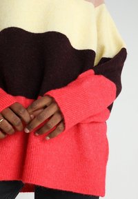 Pull tricoté coloré présentant des rayures jaunes, bordeaux et roses avec des poignets côtelés et une coupe décontractée. Texture douce et design chaud.