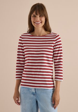 Femme souriante aux cheveux bruns portant un t-shirt à manches longues rayé rouge et blanc et un jean bleu clair, debout devant un fond beige.